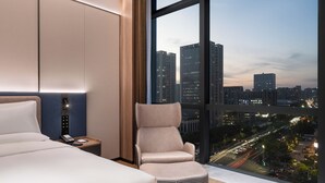 Habitación de hotel moderna con una cama grande, un escritorio con teléfono, una silla y vistas a la ciudad al anochecer.