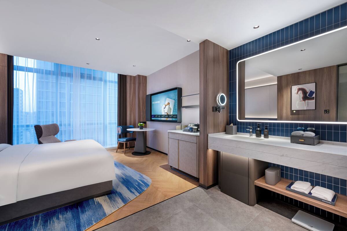 A modern hotel room with a large bed, a desk, a TV, a bathroom with a mirror, and a view of the city.