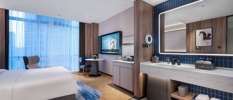 A modern hotel room with a large bed, a desk, a TV, a bathroom with a mirror, and a view of the city.