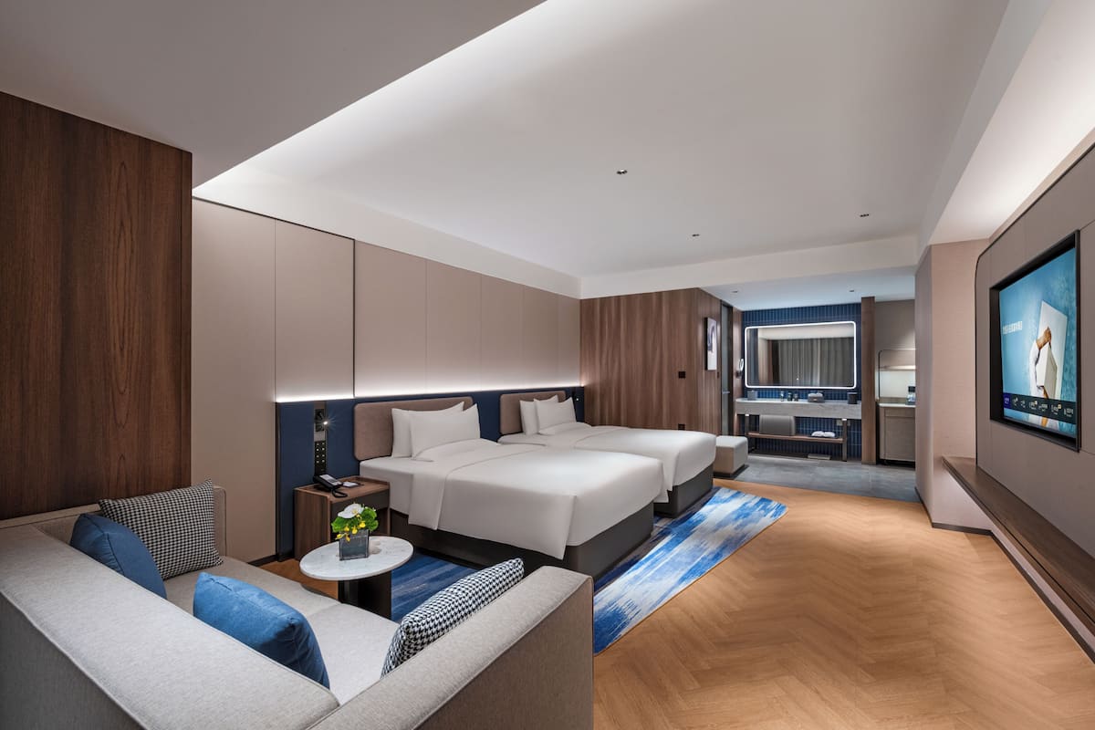 A modern hotel room with a sofa, two beds, a TV, and a kitchenette.