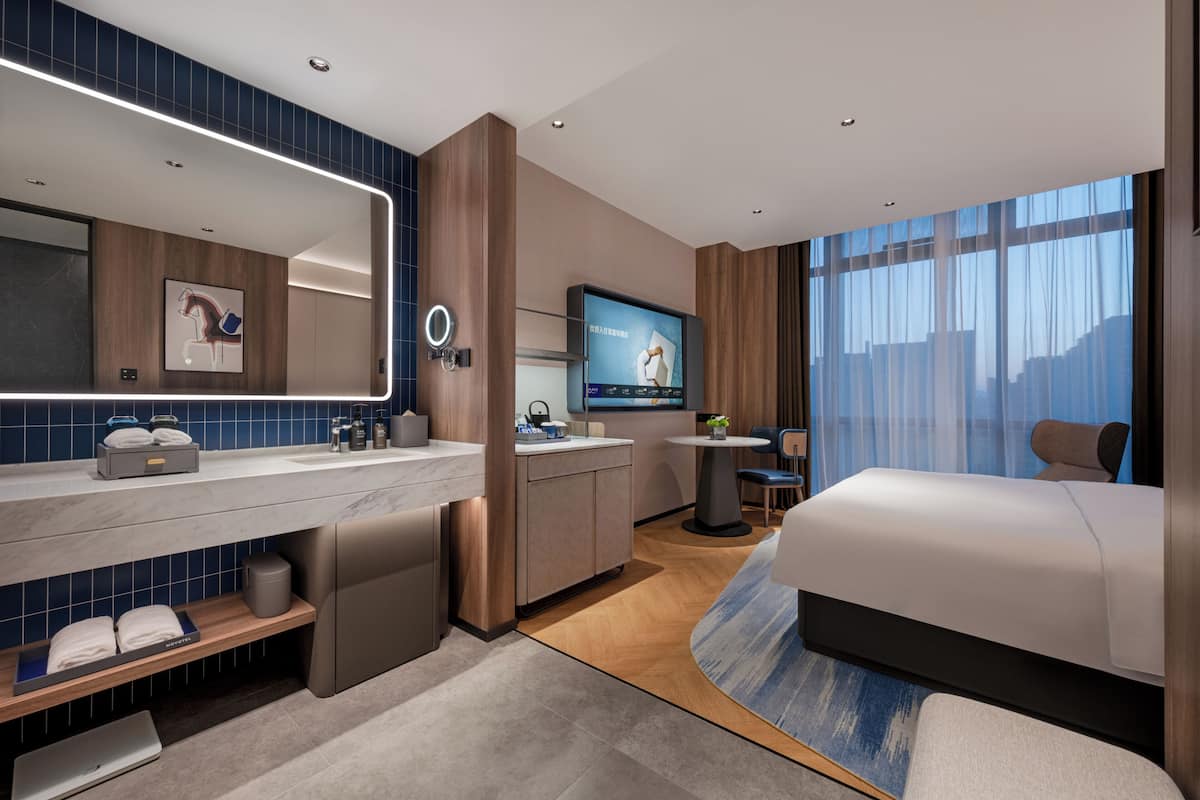 A modern hotel room with a large bed, a TV, a bathroom with a mirror, and a desk with a chair.