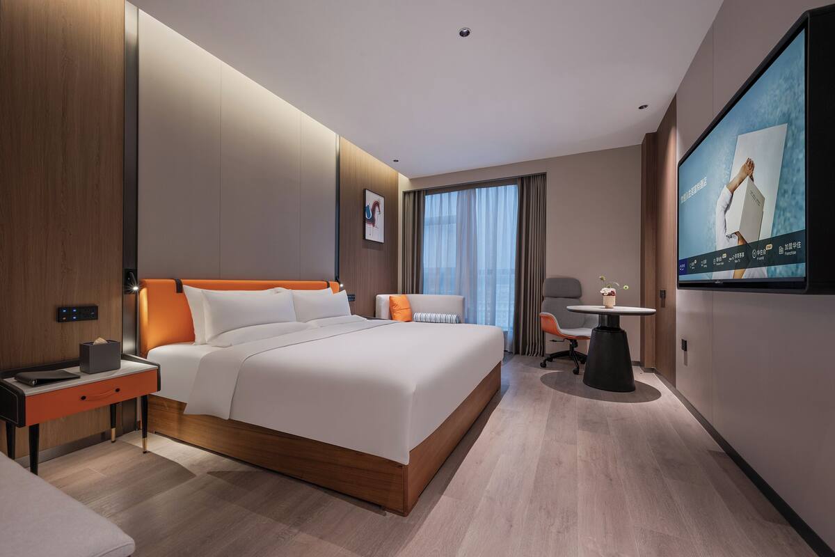 A modern hotel room with a large bed, a desk, a chair, a TV, and a window with curtains.