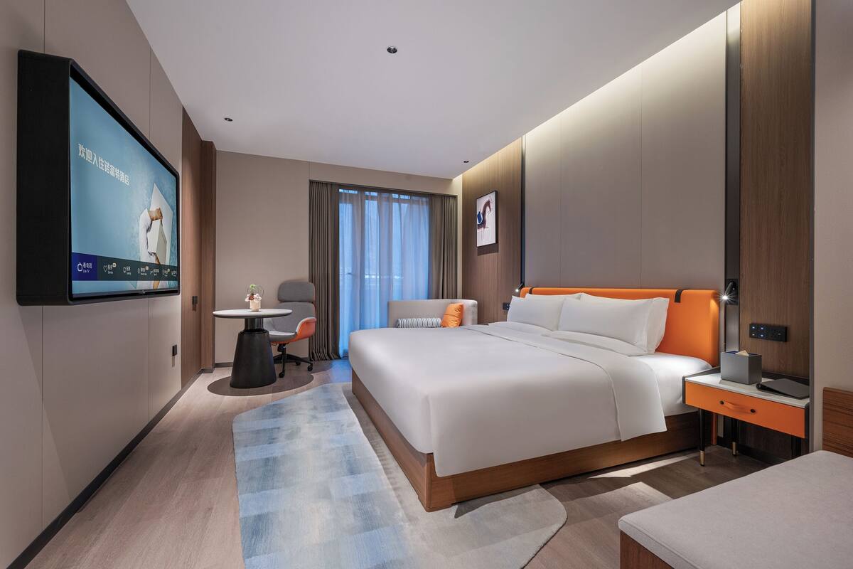 A modern hotel room with a large bed, bedside table, TV, and a desk with a chair.