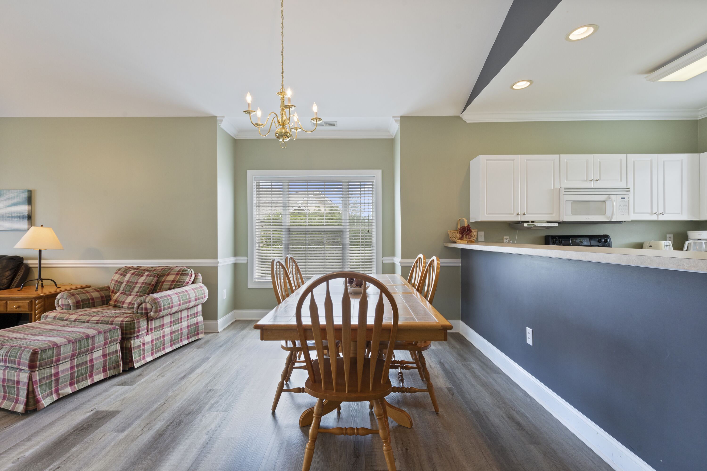 A dining area with a wooden table and chairs, a sofa, a chandelier, and a kitchen with white cabinets.