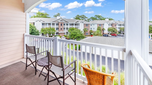 A balcony with two chairs and a wicker chair, overlooking a row of townhouses with a clear sky. - Magnolia North 305 (Myrtle Beach)