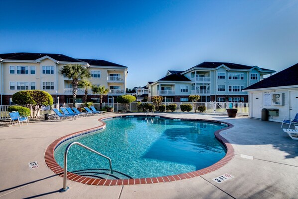 A swimming pool with a brick edge, surrounded by lounge chairs and a white fence. - Magnolia North 305 (Myrtle Beach)