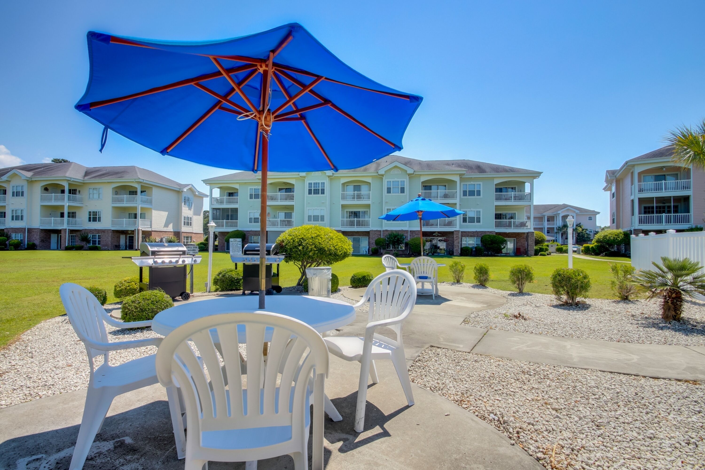 Eine Terrasse mit weißen Plastikstühlen und einem blauen Sonnenschirm, ein Grill und im Hintergrund ein Gebäude mit mehreren Balkonen.