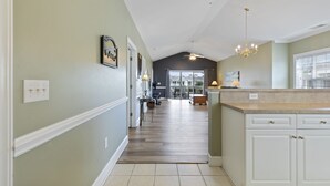 A hallway leading to a kitchen with white cabinets, a chandelier, and a dining area with a table and chairs. - Magnolia North 305 (Myrtle Beach)