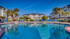 A swimming pool with lounge chairs, palm trees, and a building with balconies in the background. - Magnolia North 305 (Myrtle Beach)