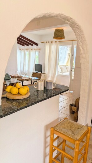 A kitchen with a stone countertop, a wicker chair, and a dining area with a table set for four. - Menorcan house with private pool, sea views, 10 minutes from the beach. (Cala en Porter)