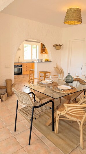 A dining area with a glass table, wicker chairs, and a kitchen in the background. - Menorcan house with private pool, sea views, 10 minutes from the beach. (Cala en Porter)