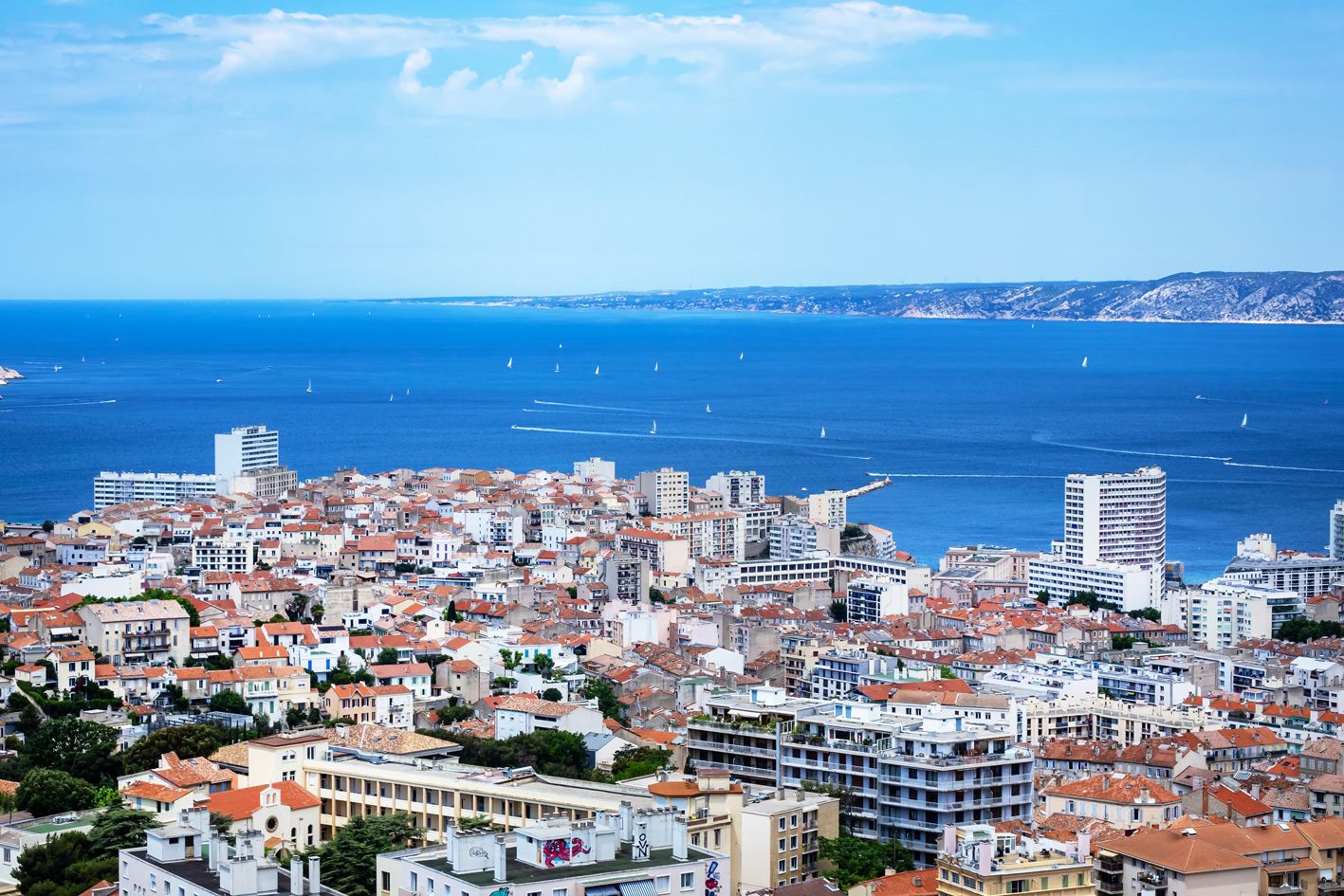 Paesaggio costiero urbano con edifici densi, un mare blu limpido e colline in lontananza.