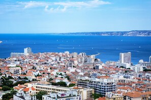 A coastal cityscape with dense urban buildings, a clear blue sea, and distant hills.