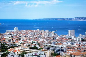 Paesaggio costiero urbano con edifici densi, un mare blu limpido e colline in lontananza.