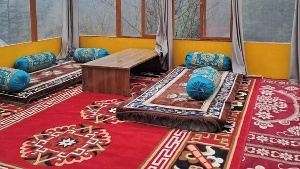 A room with a high ceiling, wooden beams, and large windows. There is a bed with patterned bedding and a small wooden table. - Thrillomania Kasol (Kasol)