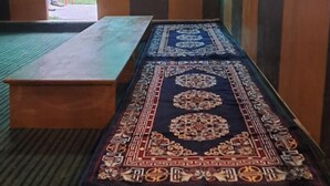 A room with a long table, a bench, and a patterned rug. There are windows with green-tinted glass. - Thrillomania Kasol (Kasol)