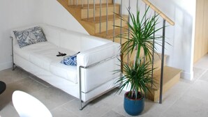 Un escalier avec des marches en bois et des rampes en métal, un canapé blanc avec un coussin à motifs, une plante bleue en pot et une chaise blanche.