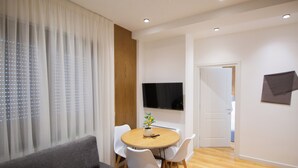 Una sala moderna con un sofá, una mesa redonda de madera con sillas, un televisor montado en la pared y una puerta que da a otra habitación.