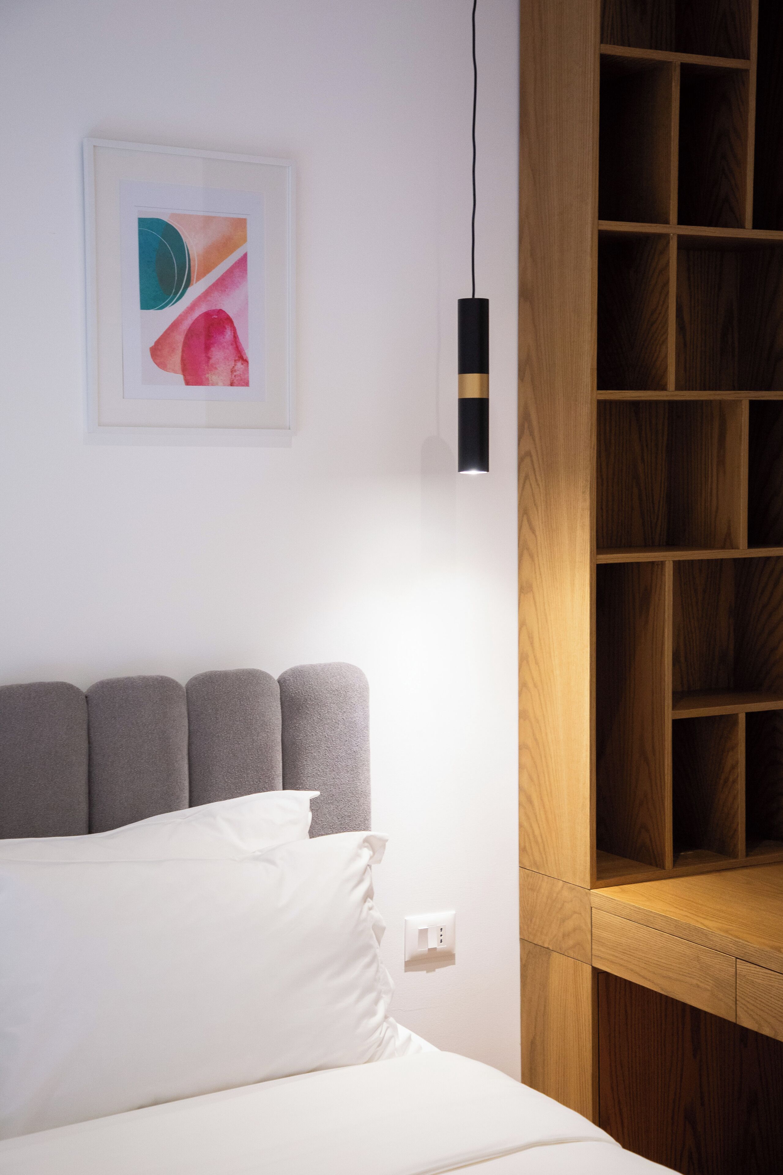 Una cama con cabecera gris, almohada blanca, luz fijada a la pared y estantería de madera.
