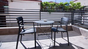 Un área de estar al aire libre con una mesa y dos sillas, una barandilla de metal y vistas a edificios y vegetación al fondo.