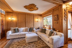 A wooden-paneled room with a large sectional sofa, wall sconces, and a view of the outdoors. - Casa Verde, with Dolomites view (Cortina d´Ampezzo)