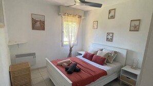 A bedroom with a bed, a ceiling fan, a window with curtains, a bedside table, and a small wooden stool. - Apt 4 pers. - swimming pool - 750 m. from the beach Private parking (Fréjus)