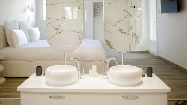A modern bathroom with two marble sinks, a large mirror, and a bed in the background.