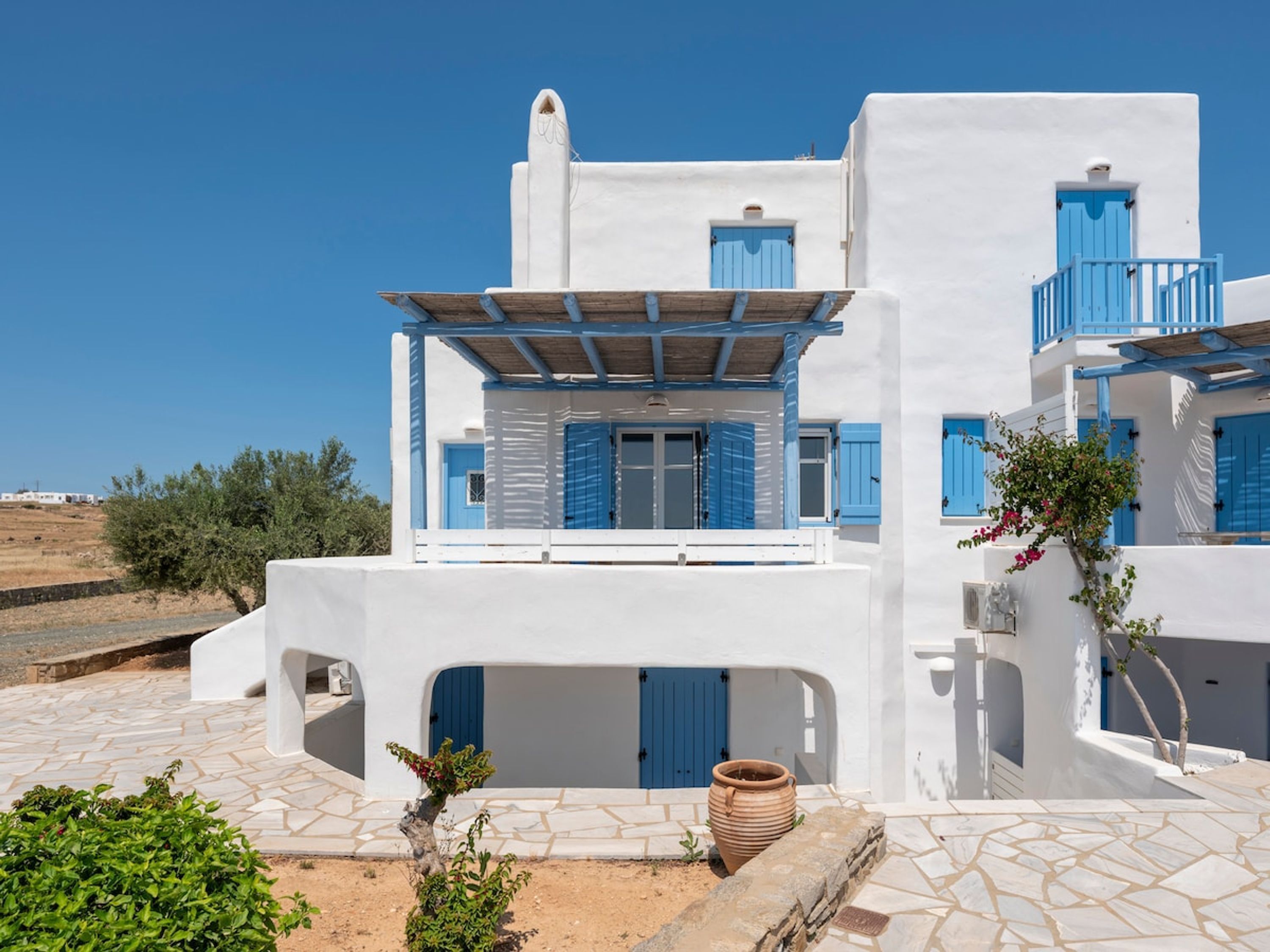 Un bâtiment blanc avec des volets bleus et un balcon, entouré de verdure et sous un ciel dégagé.
