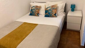 Ein ordentlich bezogenes Bett mit weißer Bettwäsche und zwei Zierkissen, ein Nachttisch mit Lampe sowie ein kleiner, weißer Schrank.