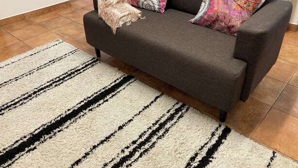Ein graues Sofa mit gemusterten Kissen, einer beigen Tagesdecke und einem Streifen Teppich in einem Wohnzimmer mit Fliesenboden.