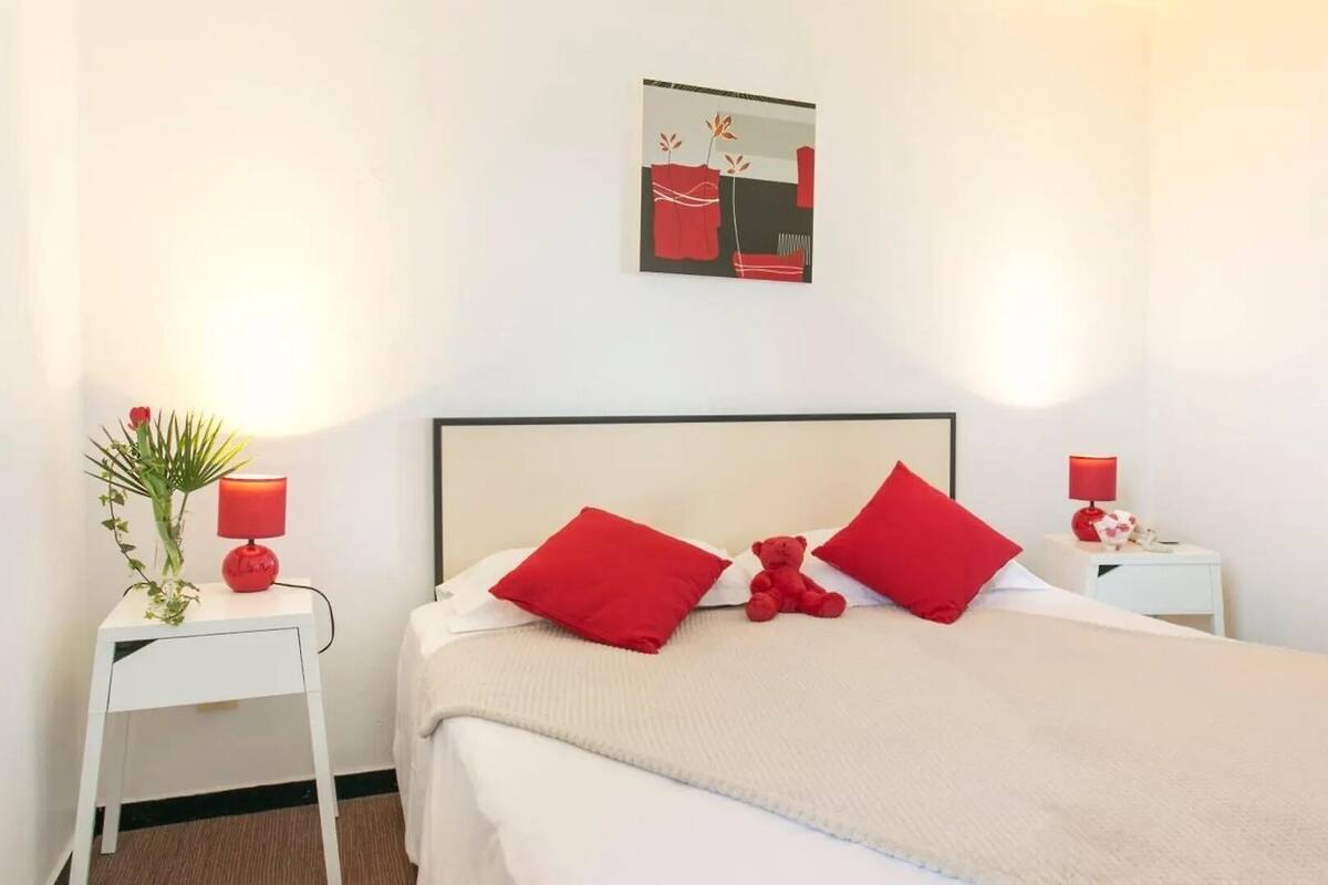 Un dormitorio con una cama, dos almohadas rojas, un osito de peluche rojo, una mesita de noche con una lámpara y un cuadro abstracto colgado en la pared.