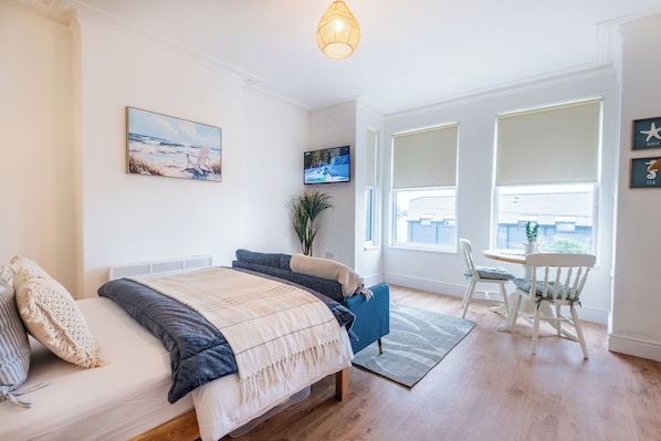 A bedroom with a bed, a TV, a plant, and a dining area with chairs.