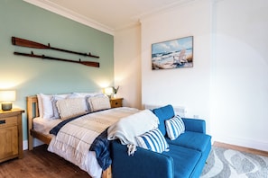 A bedroom with a bed, a blue sofa, wooden headboard, bedside tables, a painting, and a wall-mounted shelf.