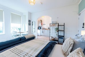 A modern bedroom with a large bed, a seating area, a kitchenette, and a view of the city.
