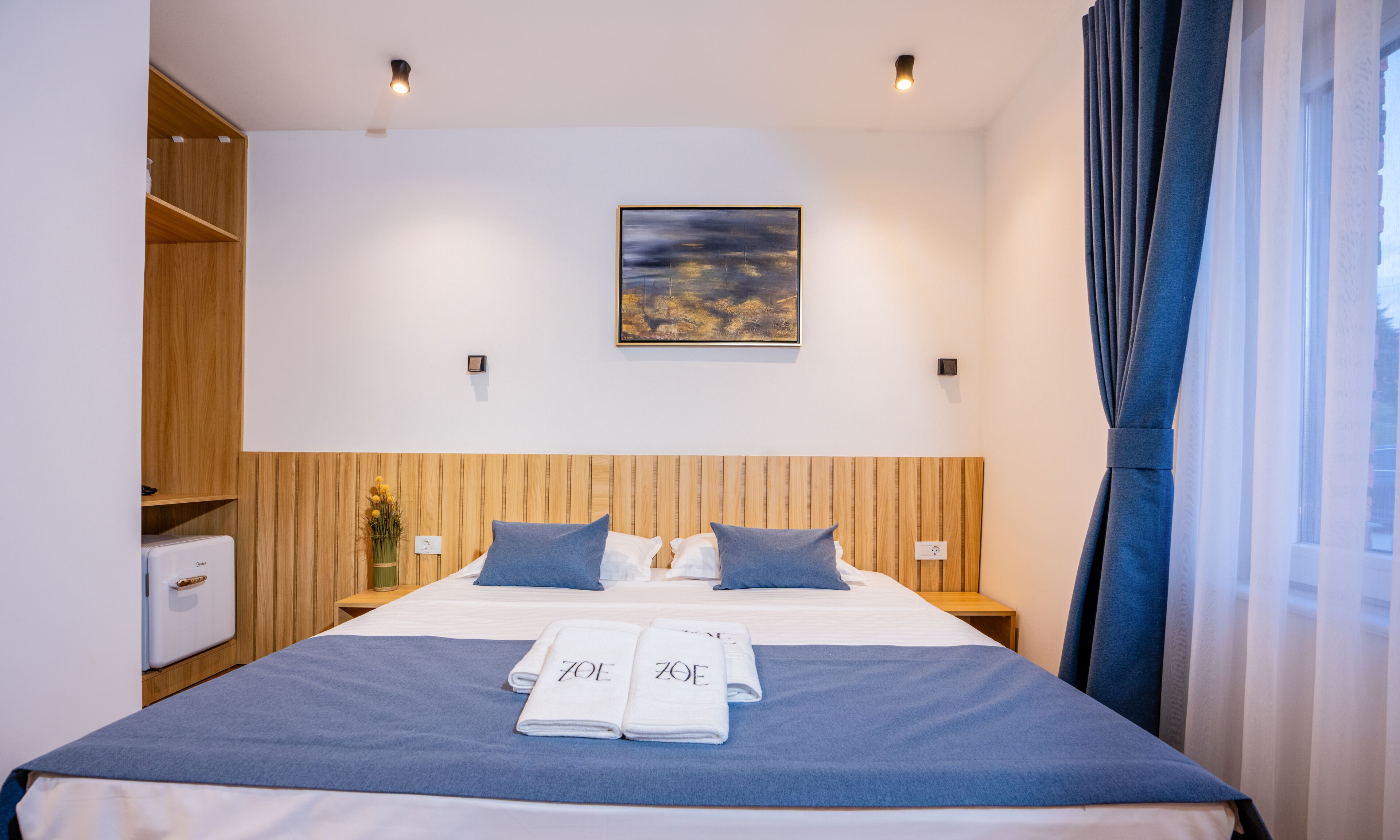 Ein Hotelzimmer mit einem Bett, einem Holz-Kopfteil, einem Bild an der Wand und einem Fenster mit Vorhängen.