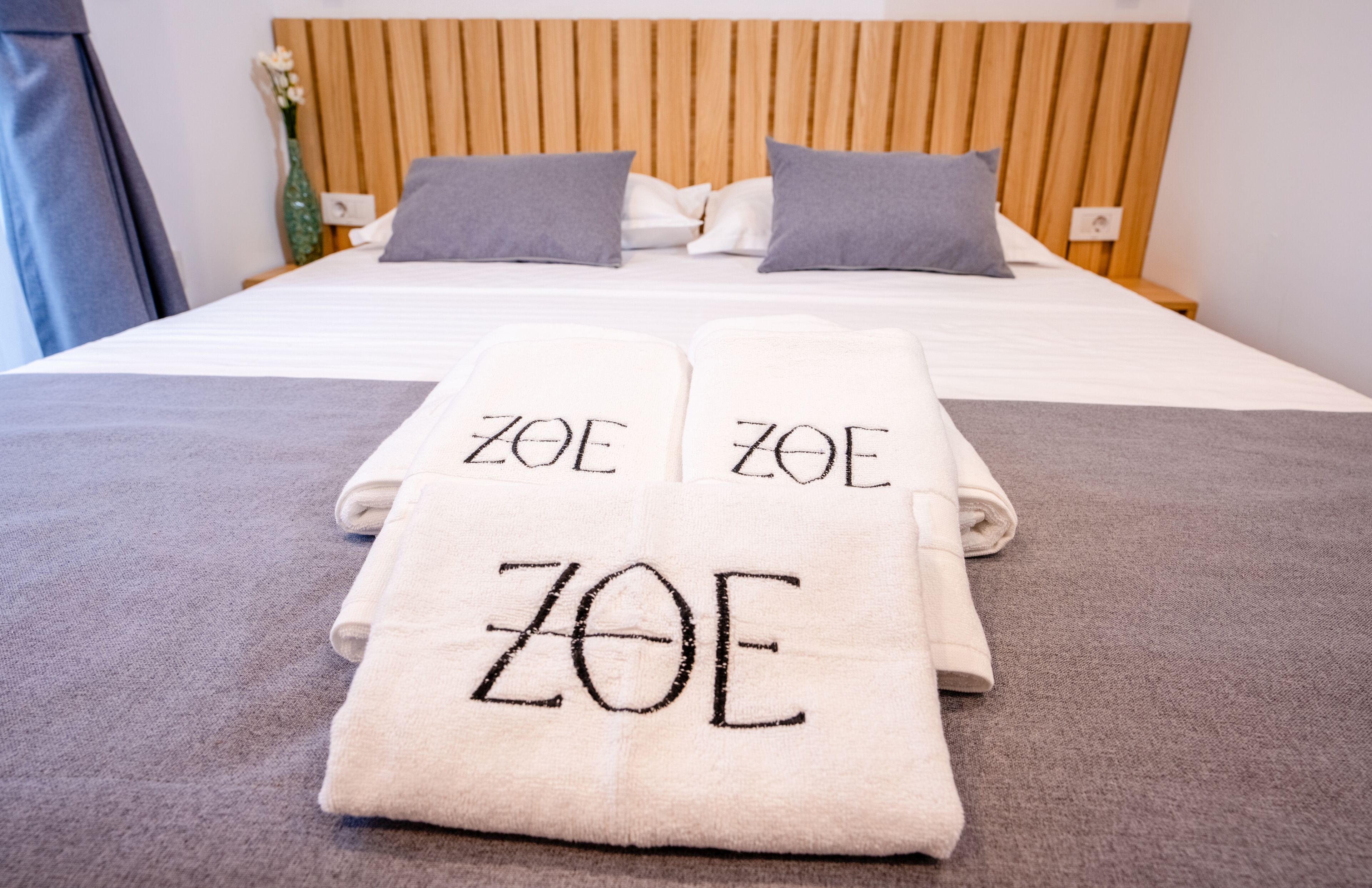 Un lit bien fait, avec des serviettes étiquetées « ZOE » et une tête de lit en bois.