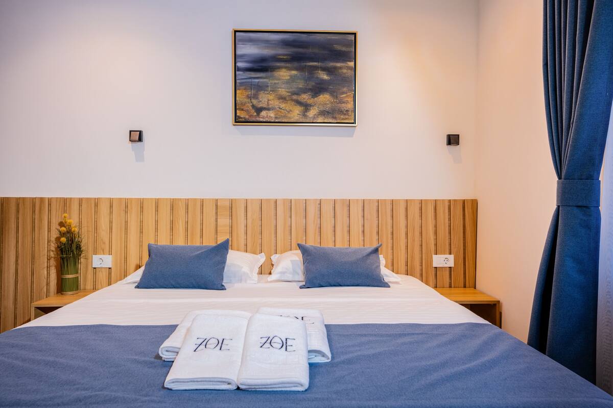 A hotel room with a wooden headboard, two pillows, a bed with a blue bedspread, two rolled towels with 'ZOE' on them, a painting on the wall, and a nightstand with a vase of flowers.