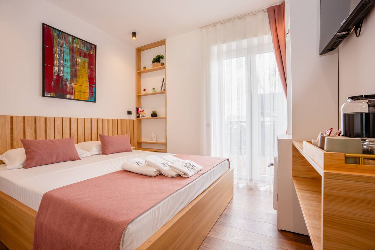 A modern hotel room with a wooden bed, a flat-screen TV, a coffee machine, and a large window with curtains.