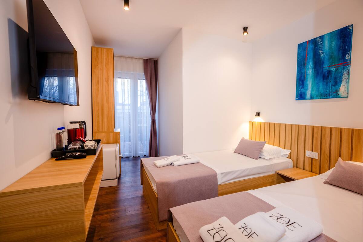 A hotel room with two beds, a TV, a wooden desk, a small table, a painting, and a window with curtains.