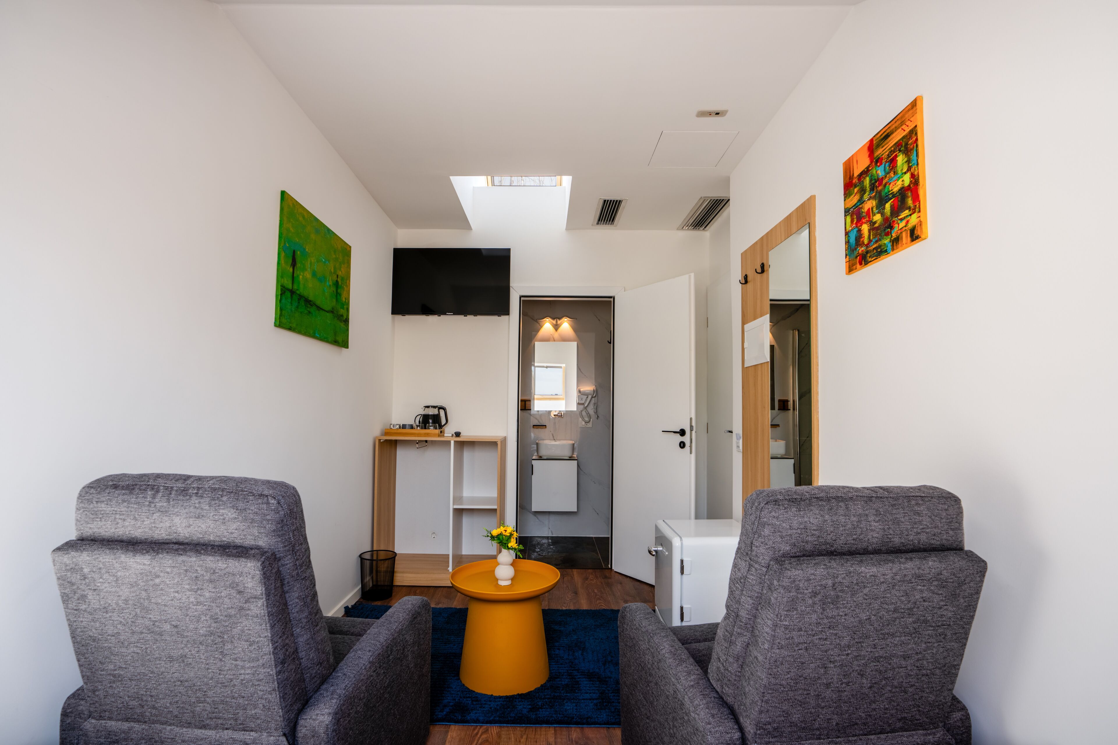 A modern room with two grey armchairs, a small orange table with a vase, a TV on the wall, and a bathroom visible through an open door.