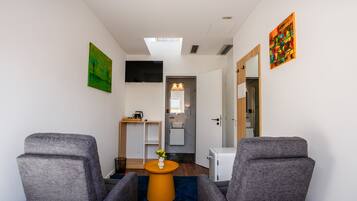 A modern room with two grey armchairs, a small orange table with a vase, a TV on the wall, and a bathroom visible through an open door.