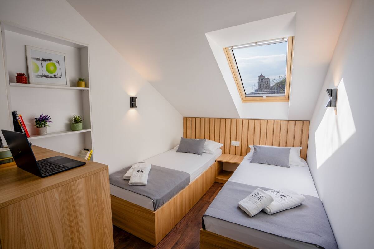 A modern, compact room with two beds, a desk, and a skylight.
