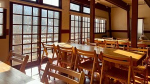 Un espace repas avec des tables et des chaises en bois, de grandes fenêtres et des poutres apparentes.