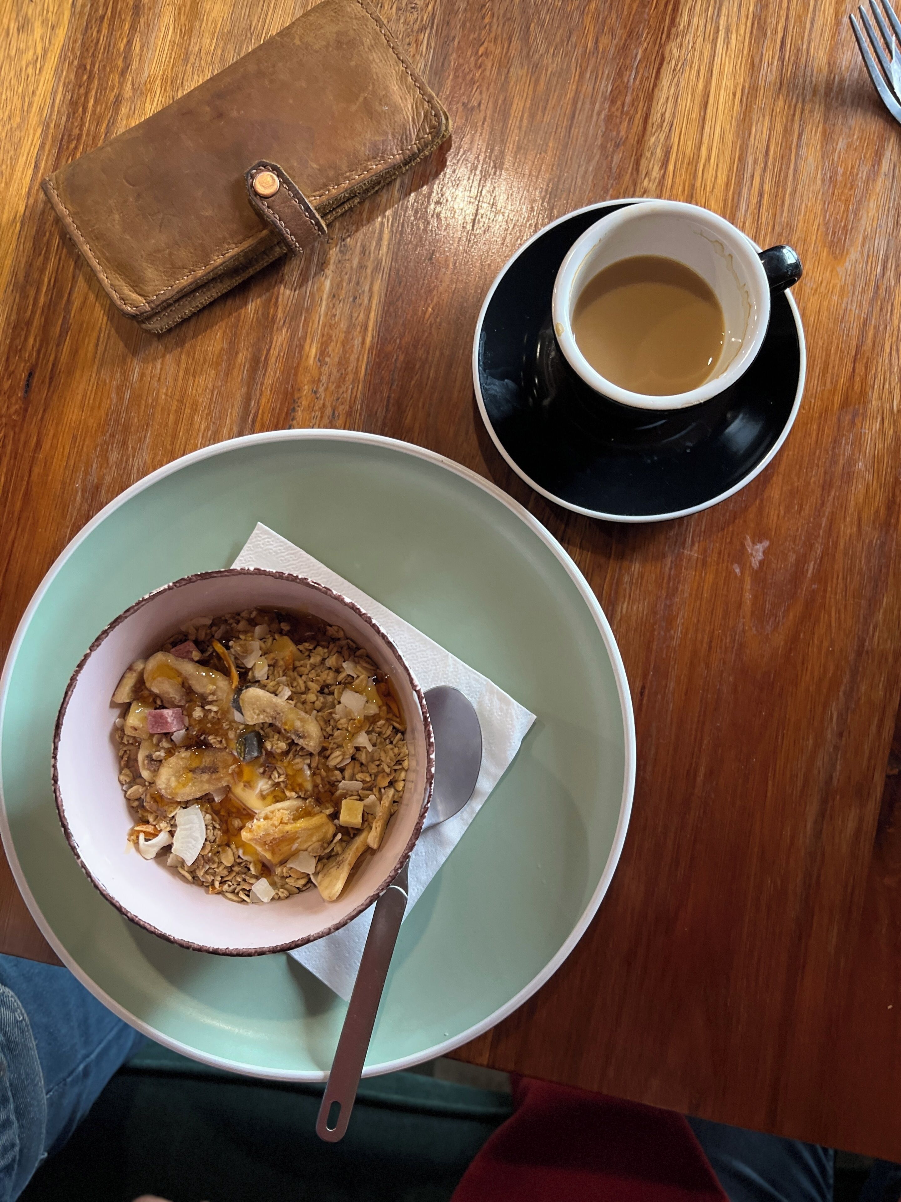 A meal with a bowl of granola and fruit, a cup of coffee, and a wallet on a wooden table.