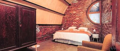 A bedroom with a brick wall, a round window, a bed, a wardrobe, and a chair.