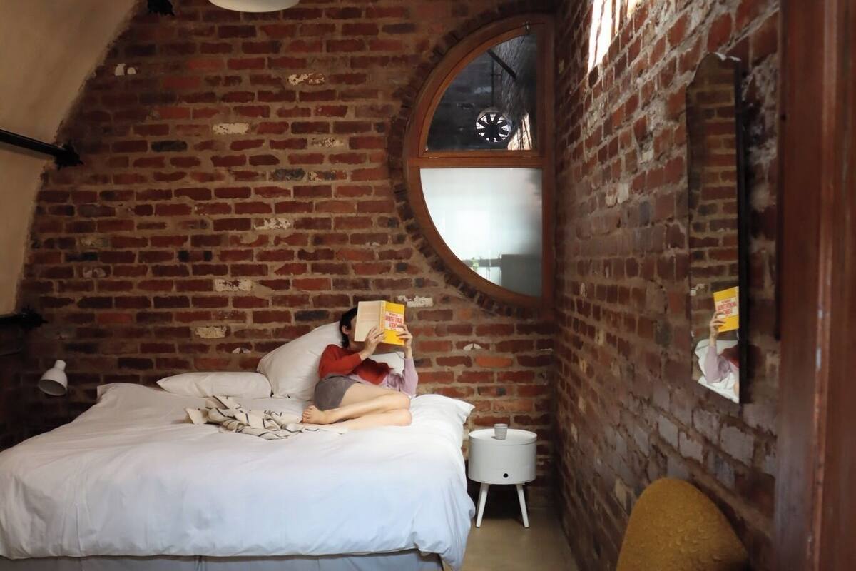 A bedroom with a brick wall, a round mirror, a bed, a nightstand, and a chair.