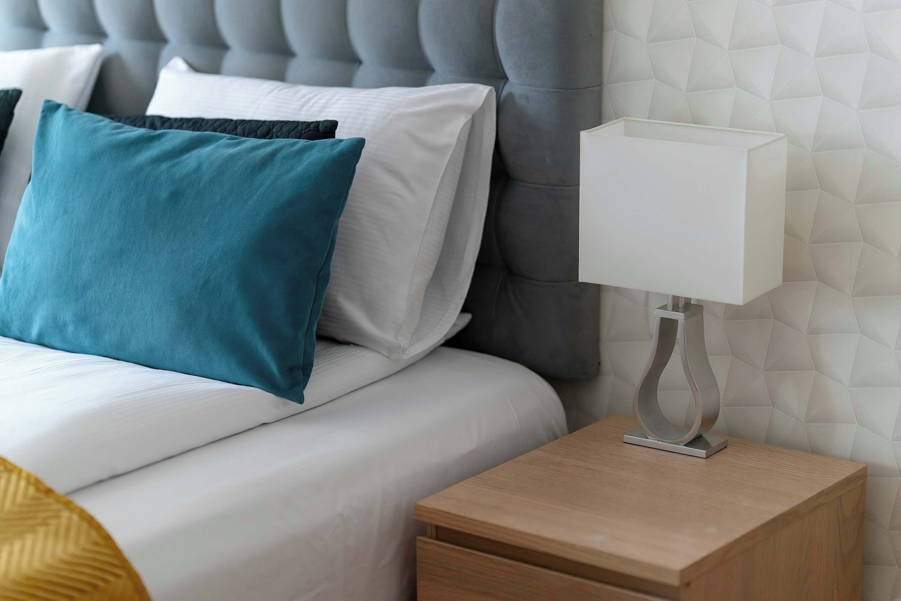 Een net opgemaakt bed met witte en teal kussens, een grijs gestoffeerd hoofdbord, een nachtkastje met een witte lamp en een houten nachtkastje.