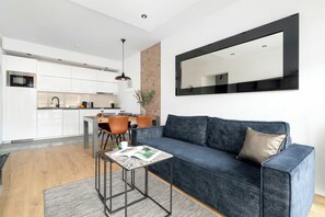 Ein zeitgemäßer Wohnraum mit einem dunkelblauen Sofa, einem kleinen Tisch mit einer Zeitschrift, einer Küchenzeile mit weißen Schränken und einem großen Spiegel an der Wand.
