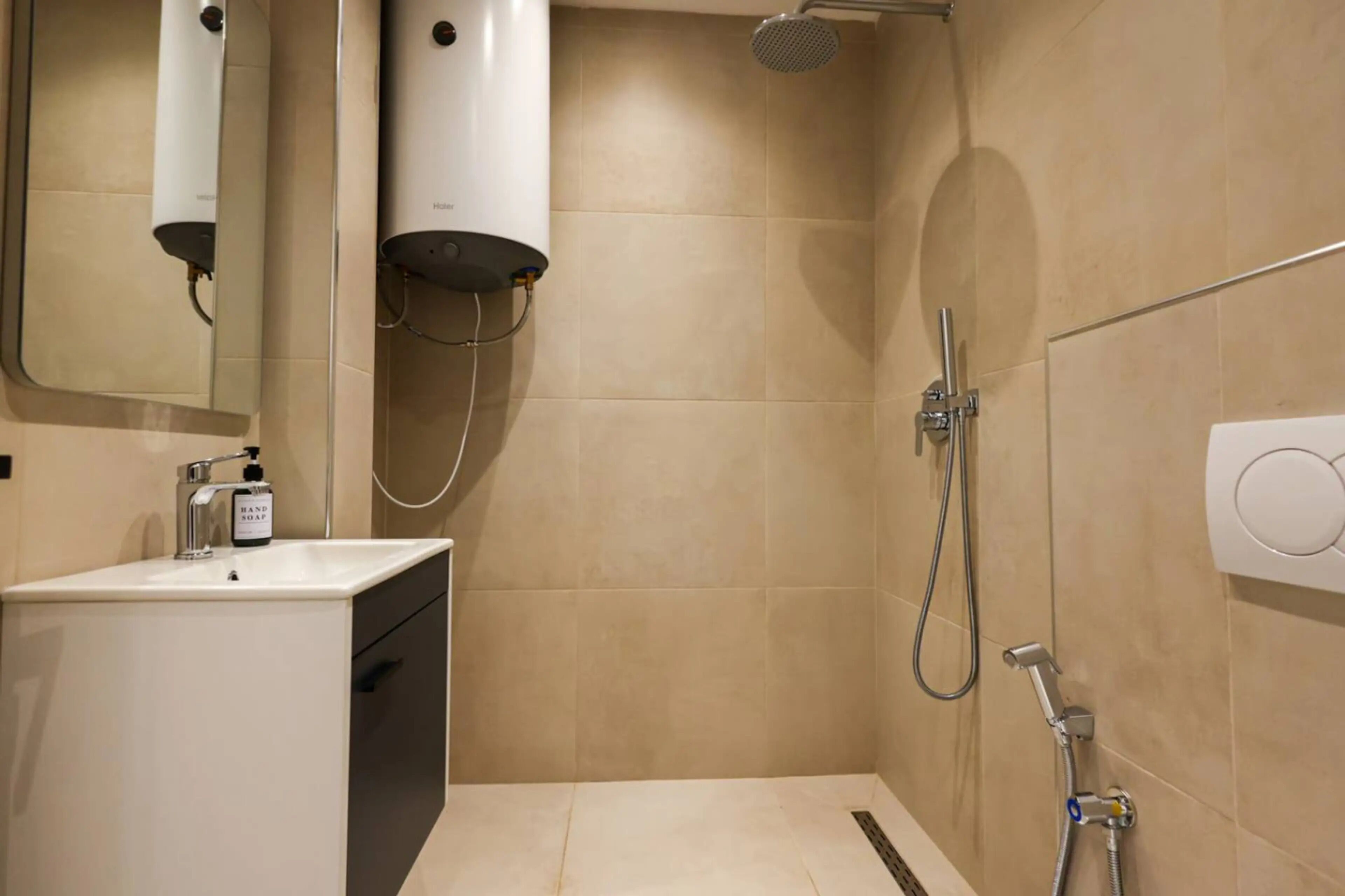 Une salle de bain d’hôtel équipée d’un lavabo, d’un distributeur de savon liquide, d’un miroir, d’une douche avec un pommeau de douche à main et d’un panneau de commande fixé au mur.