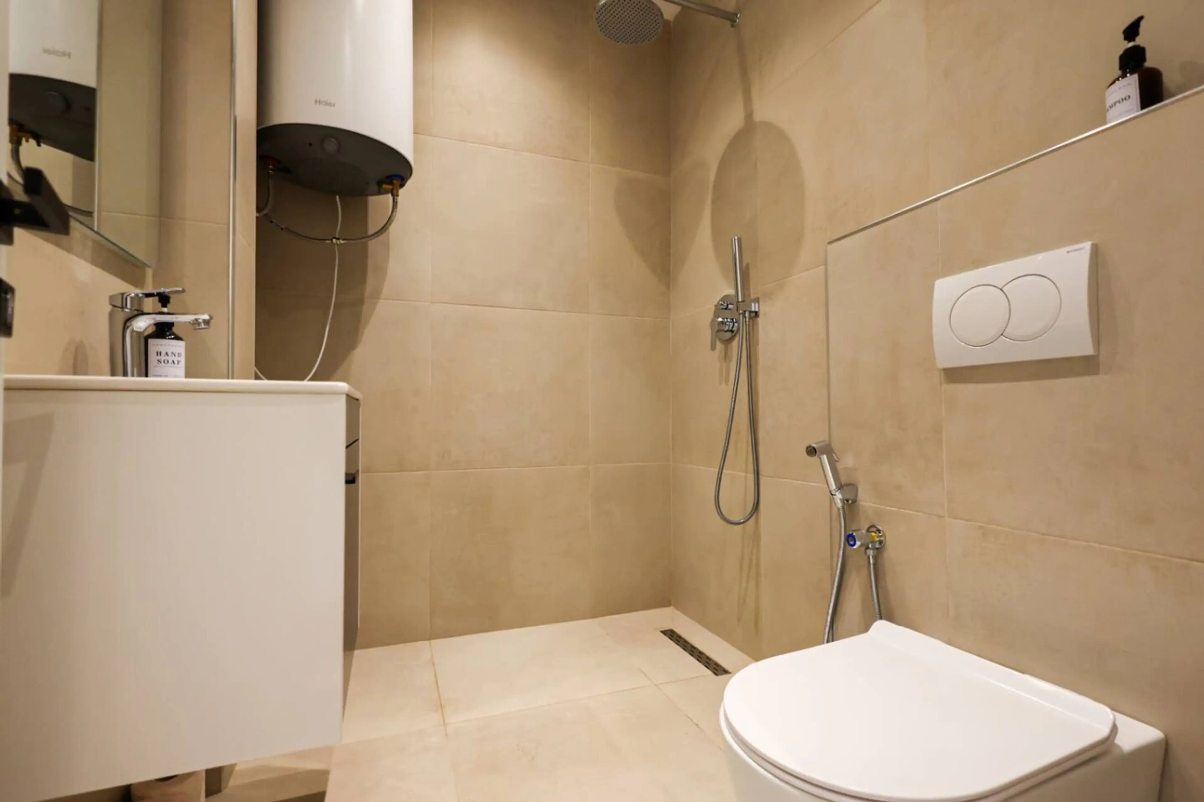 A modern bathroom with a toilet, a hand soap dispenser, a shower with a handheld showerhead, and a wall-mounted control panel.